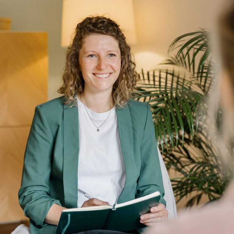Junge Frau mit lockigem Haar, lächelt, trägt einen Blazer und eine weiße Bluse.